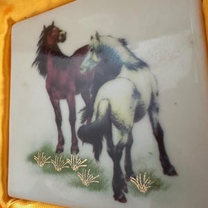 May include: A white ceramic tile with a painted image of two horses. The horse on the left is brown with a black mane and tail. The horse on the right is white with a black mane and tail. Both horses are standing in a field of green grass with gold accents.