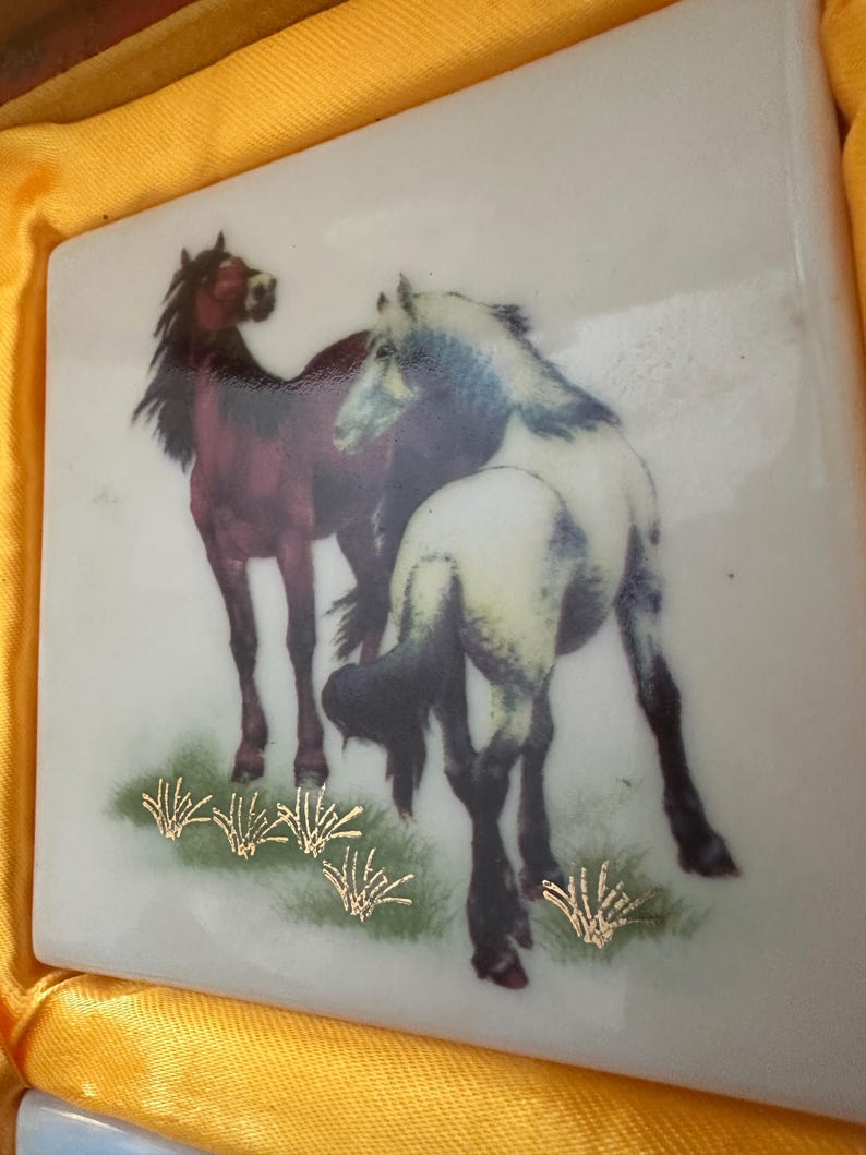 May include: A white ceramic tile with a painted image of two horses. The horse on the left is brown with a black mane and tail. The horse on the right is white with a black mane and tail. Both horses are standing in a field of green grass with gold accents.
