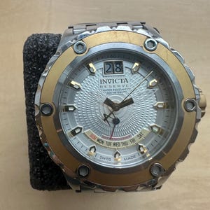 May include: A gold and silver Invicta Reserve watch with a white face and a day-date retrograde display. The watch is water resistant to 500 meters.