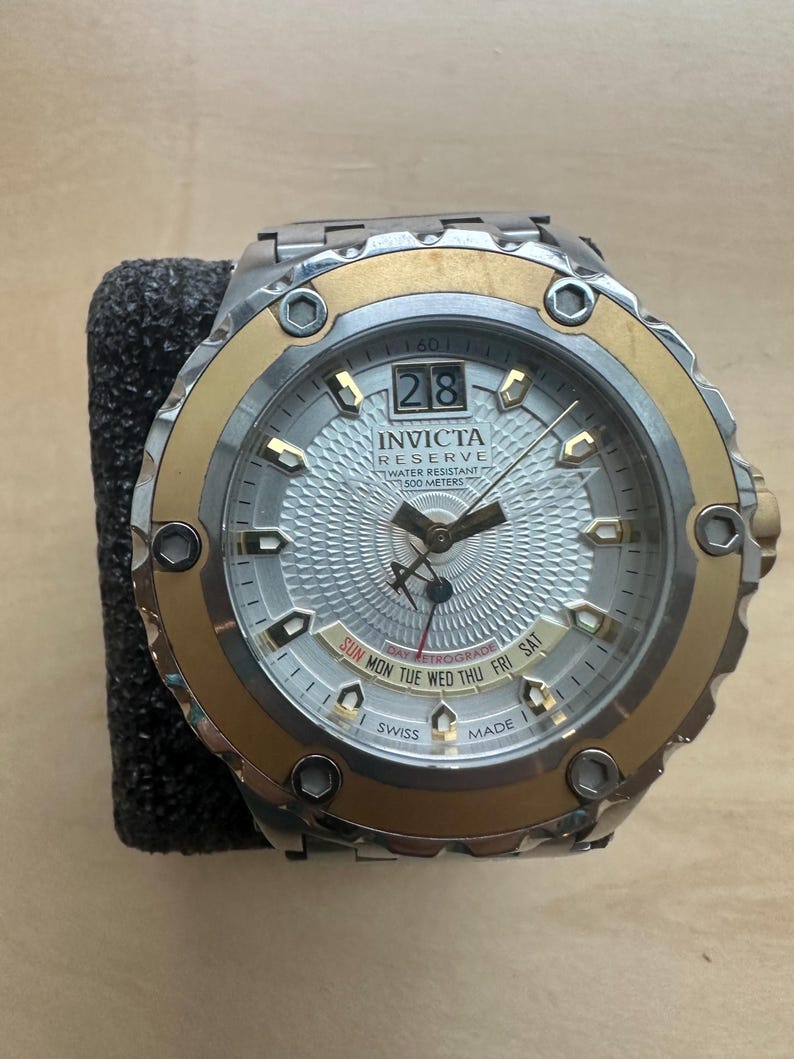 May include: A gold and silver Invicta Reserve watch with a white face and a day-date retrograde display. The watch is water resistant to 500 meters.