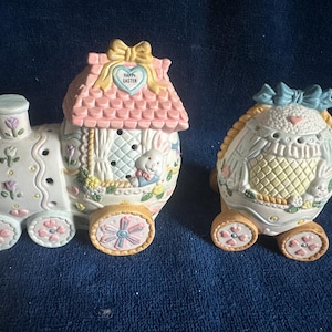 May include: A set of two ceramic Easter train cars. The first car is a house with a pink roof and the text "Happy Easter" on the front. The second car is an egg with a blue bow. Both cars have wheels and are decorated with flowers and bunnies.
