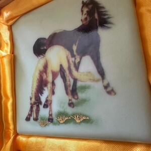 May include: A white ceramic tile with a painted image of two brown horses in a grassy field. The horses are facing different directions, and the one in the foreground is partially obscured by the other horse. The tile has a gold border and a gold design around the horses.
