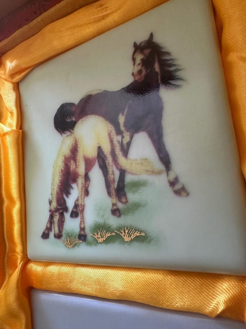May include: A white ceramic tile with a painted image of two brown horses in a grassy field. The horses are facing different directions, and the one in the foreground is partially obscured by the other horse. The tile has a gold border and a gold design around the horses.