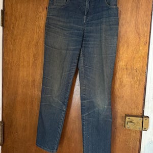 Op de afbeelding: Een blauwe spijkerbroek hangt aan een metalen hanger. De jeans heeft een klassiek ontwerp met vijf zakken, een knoop- en ritssluiting en een vervaagde wassing. De jeans heeft een rechte pijp en lijkt een middelhoge taille te hebben.