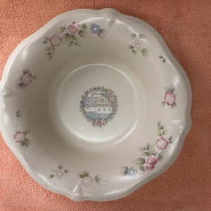 Op de afbeelding: Een witte keramische kom met een gegolfde rand en een bloemenontwerp. De kom heeft een centraal beeld van een huis met een krans en bloemen.