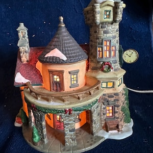May include: A ceramic Christmas village house with a stone tower and a round building. The house has a red roof, a clock, and a green garland. The windows are lit, and there is a small amount of snow on the roof.