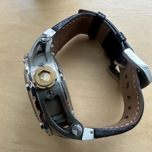 May include: A silver and black wristwatch with a black leather band. The watch has a large, round face with a gold-colored crown and a small, gold-colored detail on the face.