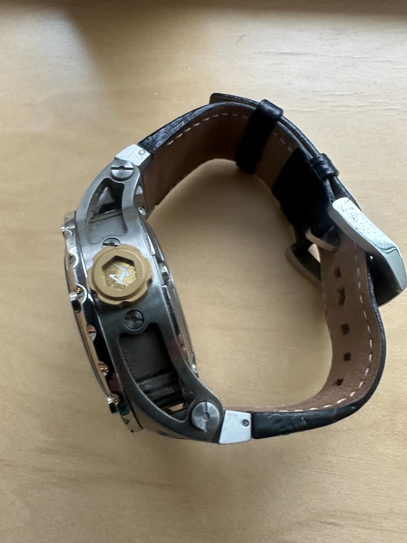 May include: A silver and black wristwatch with a black leather band. The watch has a large, round face with a gold-colored crown and a small, gold-colored detail on the face.