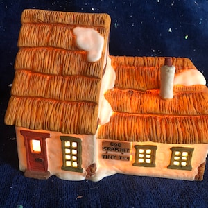 Puede incluir: Una casa de cerámica de un pueblo navideño con tejado de paja, detalles de nieve y ventanas iluminadas. El letrero dice "Bob Crachit Tiny Tim". La casa tiene una chimenea y emite una luz cálida y acogedora.