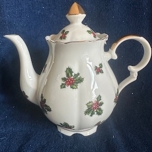 Puede incluir: Tetera de porcelana blanca con ribete dorado y diseño de acebo. La tetera tiene una tapa con un pomo dorado y un asa.