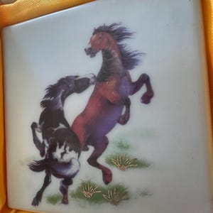 May include: Two horses, one brown and one black and white, are depicted in a playful pose on a white ceramic tile. The tile has a light blue background and gold accents.
