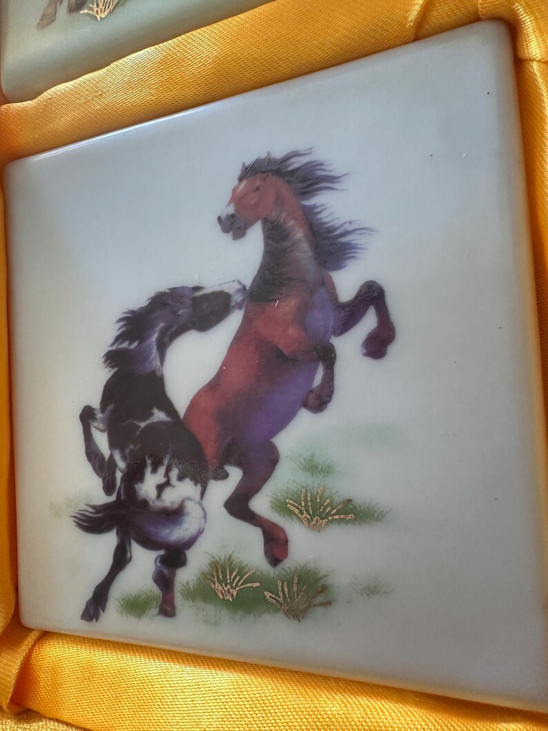 May include: Two horses, one brown and one black and white, are depicted in a playful pose on a white ceramic tile. The tile has a light blue background and gold accents.