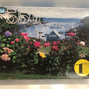 May include: A 550-piece jigsaw puzzle depicting a colourful scene of a harbour with boats, a house, and a lush garden with pink, purple, and white flowers. The box includes a voucher for £1 off.