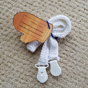 May include: White mitten clips with a mitten-shaped tag. The tag reads "Mitten Clips" and "Created with love by NanaInDoor". The clips have white plastic clasps. The clips are made of a white, knitted material.