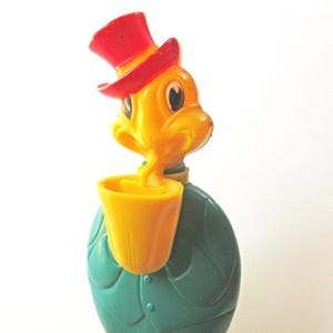 May include: A vintage plastic bottle shaped like a character wearing a red top hat. The character has a yellow head and is holding a yellow cup to its mouth. The body of the bottle is teal and molded to resemble a jacket with visible buttons.