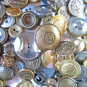 May include: A collection of assorted metal buttons in various sizes and designs. The buttons are in shades of silver, gold, and bronze, with details like rope edges, embossed patterns, and two or four-hole configurations. Some buttons have a shiny, reflective surface.