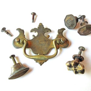 May include: An assortment of antique brass hardware, including a decorative handle and several knobs with screws. The handle has a curved design with ornate detailing. The knobs feature various shapes and floral patterns, all with a vintage aesthetic.