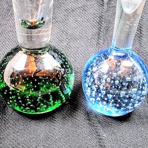 May include: Two glass bottle stoppers, one green and one blue, each with a round, bubble-filled base and a clear stem. The green stopper has a vibrant, emerald hue, while the blue one is a lighter, sky blue. Both are on a dark gray surface.