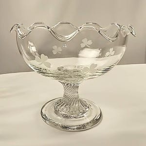 May include: A clear glass footed bowl with a scalloped rim and etched shamrock designs. The bowl has a twisted stem and a round base. The bowl is likely used for serving or display.