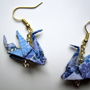 Puede incluir: Un par de pendientes de grulla de origami azul con ganchos dorados y cuentas transparentes. Las grullas est&aacute;n hechas de papel estampado con detalles dorados.