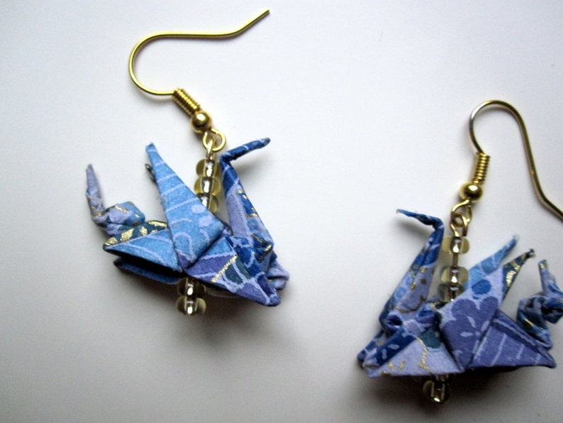 Puede incluir: Un par de pendientes de grulla de origami azul con ganchos dorados y cuentas transparentes. Las grullas est&aacute;n hechas de papel estampado con detalles dorados.