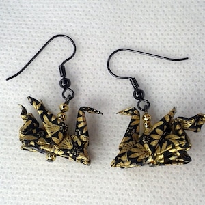Puede incluir: Un par de pendientes de grulla de origami dorada y negra con ganchos de metal negro. Las grullas est&aacute;n hechas de tela con un patr&oacute;n floral dorado y negro.
