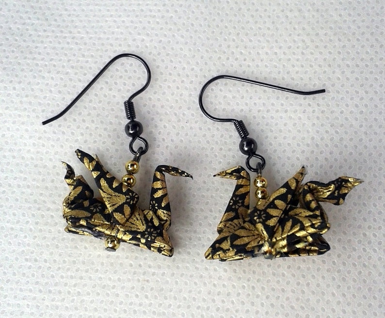 Puede incluir: Un par de pendientes de grulla de origami dorada y negra con ganchos de metal negro. Las grullas est&aacute;n hechas de tela con un patr&oacute;n floral dorado y negro.