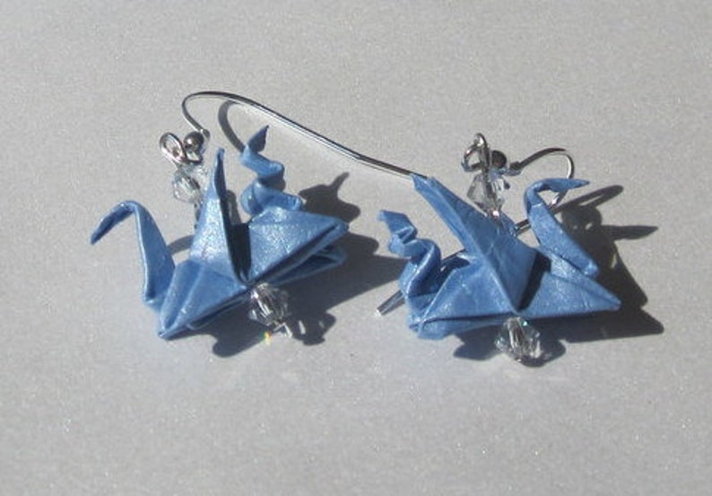 Puede incluir: Un par de pendientes de grulla de origami azul con ganchos plateados y detalles de cristal.