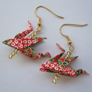 Puede incluir: Un par de pendientes de grulla origami roja y dorada con cuentas blancas. Las grullas est&aacute;n hechas de tela estampada y tienen detalles dorados.