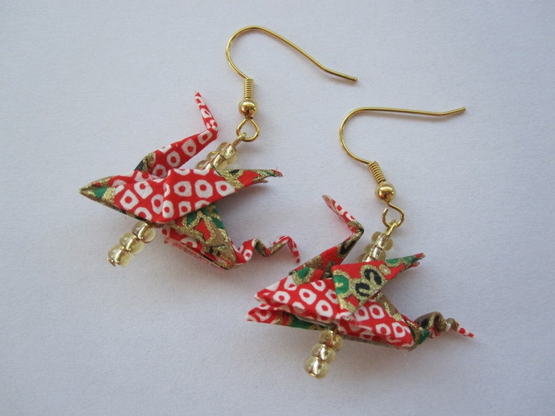 Puede incluir: Un par de pendientes de grulla origami roja y dorada con cuentas blancas. Las grullas est&aacute;n hechas de tela estampada y tienen detalles dorados.