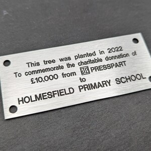 May include: A silver metal plaque with black text that reads: "This tree was planted in 2022 To commemorate the charitable donation of &pound;10,000 from PRESSPART to HOLMESFIELD PRIMARY SCHOOL."