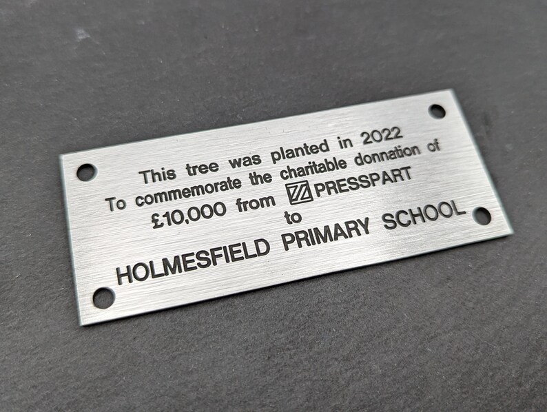 May include: A silver metal plaque with black text that reads: "This tree was planted in 2022 To commemorate the charitable donation of &pound;10,000 from PRESSPART to HOLMESFIELD PRIMARY SCHOOL."