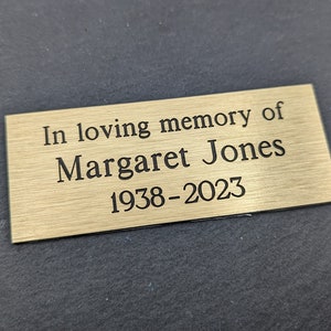 May include: A gold-colored metal plaque with the inscription "In loving memory of Margaret Jones 1938-2023".