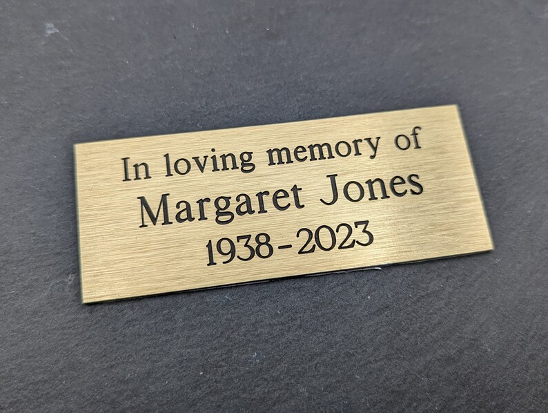 May include: A gold-colored metal plaque with the inscription "In loving memory of Margaret Jones 1938-2023".
