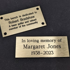 May include: Two gold-colored metal plaques with engraved text. The top plaque reads "This bench is dedicated to Robert Bradshaw A keen golfer and valued member of the community". The bottom plaque reads "In loving memory of Margaret Jones 1938-2023".