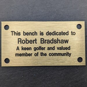 May include: A gold-colored metal plaque with rounded corners and four holes. The plaque has the following text: "This bench is dedicated to Robert Bradshaw A keen golfer and valued member of the community"