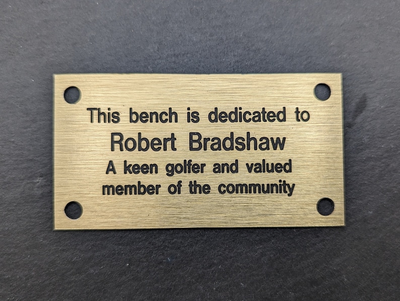 May include: A gold-colored metal plaque with rounded corners and four holes. The plaque has the following text: "This bench is dedicated to Robert Bradshaw A keen golfer and valued member of the community"