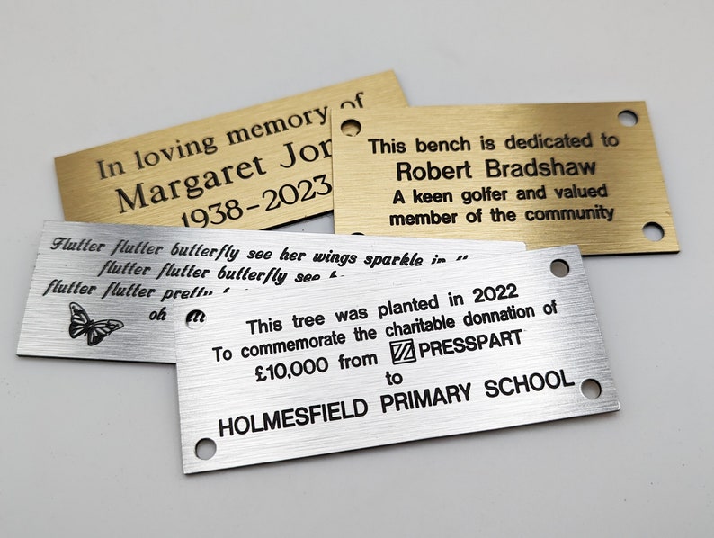 May include: Three engraved metal plaques with different messages. The top plaque is gold and reads "In loving memory of Margaret Jor 1938-2023". The middle plaque is silver and reads "Flutter flutter butterfly see her wings sparkle in... flutter flutter butterfly see... flutter flutter preff... oh". The bottom plaque is silver and reads "This tree was planted in 2022 To commemorate the charitable donation of &pound;10,000 from PRESSPART to HOLMESFIELD PRIMARY SCHOOL".