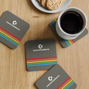 Może przedstawiać: Cztery kwadratowe, szare podstawki z logo Commodore i tęczowym wzorem w paski. Na jednej podstawce stoi biały kubek z kawą, a obok talerz z ciasteczkami. Podstawki leżą na jasnej drewnianej powierzchni.