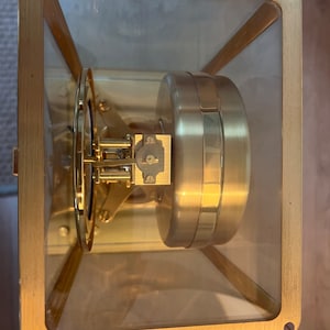 K&ouml;nnte beinhalten: Ein vergoldeter Uhrwerk, das in einem Rahmen aus Glas und vergoldetem Metall eingefasst ist. Das Zifferblatt der Uhr ist durch das Glas sichtbar.