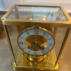 K&ouml;nnte beinhalten: Eine goldfarbene, gl&auml;serne Analoguhr mit wei&szlig;em Zifferblatt und schwarzen Zeigern. Die Uhr ist mit "Atmos" und "Jaeger-LeCoultre" beschriftet.
