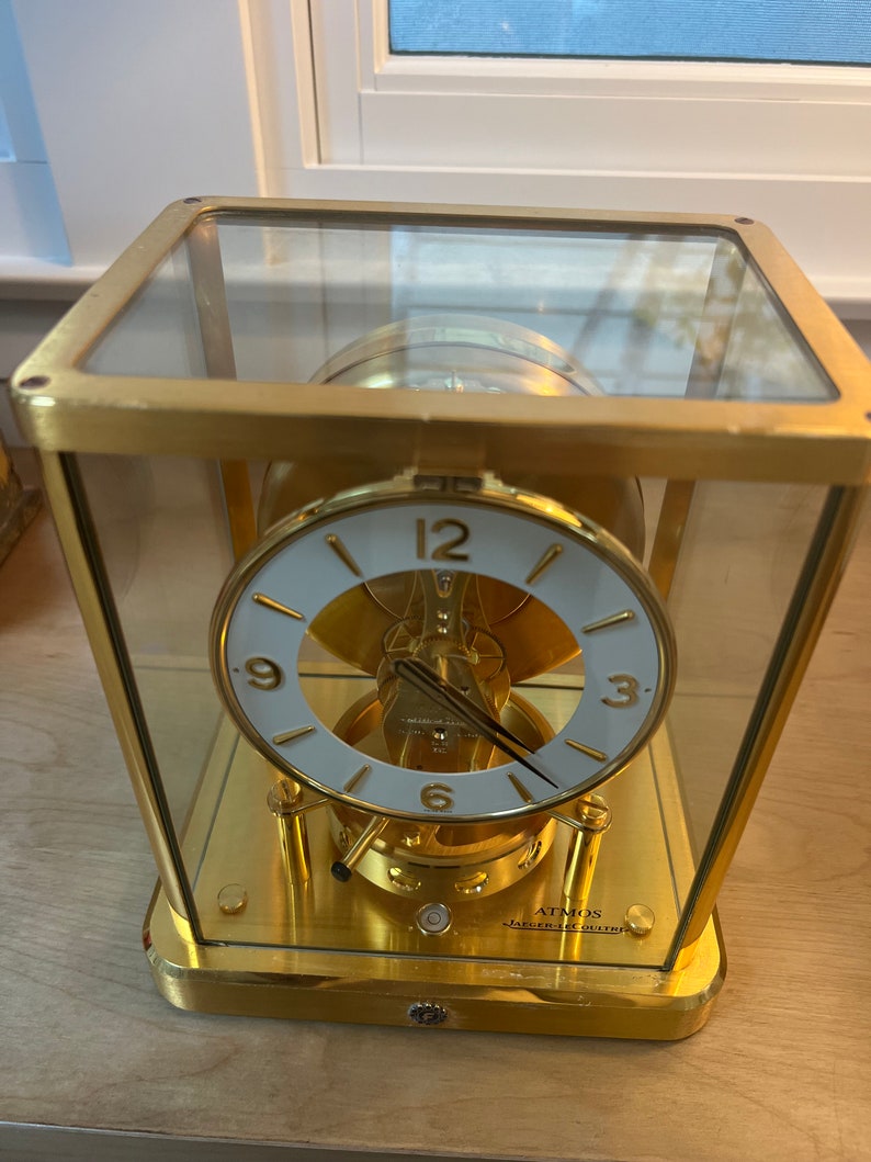 K&ouml;nnte beinhalten: Eine goldfarbene, gl&auml;serne Analoguhr mit wei&szlig;em Zifferblatt und schwarzen Zeigern. Die Uhr ist mit "Atmos" und "Jaeger-LeCoultre" beschriftet.