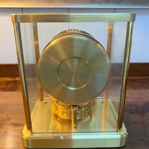 K&ouml;nnte beinhalten: Eine goldfarbene Uhr mit einem runden Zifferblatt und einem Glasgeh&auml;use. Die Uhr steht auf einer Holzoberfl&auml;che. Der Text "Corbeta "Florentina" Capt. D. Manuel Ferrer" ist auf der Oberfl&auml;che hinter der Uhr sichtbar.