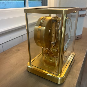 K&ouml;nnte beinhalten: Eine goldfarbene Uhr mit einem Glasgeh&auml;use, das die komplizierten inneren Mechanismen des Uhrwerks zeigt. Das Zifferblatt der Uhr hat r&ouml;mische Ziffern und einen wei&szlig;en Hintergrund.