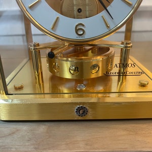 K&ouml;nnte beinhalten: Eine goldfarbene Atmos-Uhr von Jaeger-LeCoultre. Das Zifferblatt der Uhr ist wei&szlig; mit schwarzen Zeigern und r&ouml;mischen Ziffern. Die Uhr wird durch Temperaturschwankungen in der Umgebung angetrieben. Die Uhr steht auf einer Holzoberfl&auml;che.