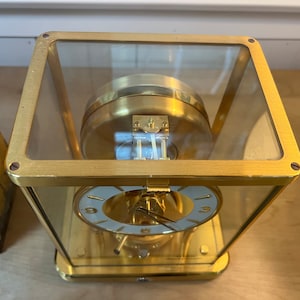 K&ouml;nnte beinhalten: Eine goldfarbene Uhr mit einem Glasgeh&auml;use und einem wei&szlig;en Zifferblatt. Die Uhr hat r&ouml;mische Ziffern f&uuml;r die Stunden und einen Sekundenzeiger. Die Uhr steht auf einer Holzoberfl&auml;che.