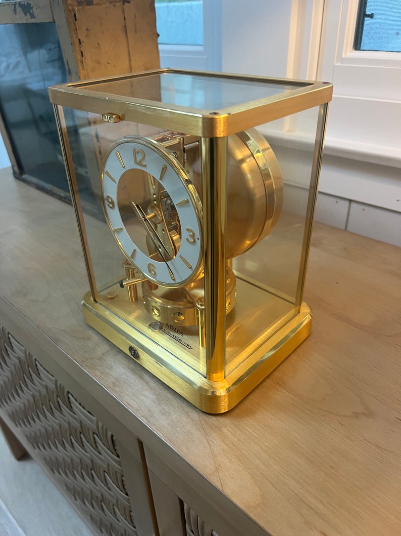 K&ouml;nnte beinhalten: Eine goldfarbene Uhr mit wei&szlig;em Zifferblatt und schwarzen r&ouml;mischen Ziffern. Die Uhr ist in einer Glaskuppel eingeschlossen und steht auf einer Holzoberfl&auml;che.