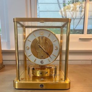 K&ouml;nnte beinhalten: Eine goldfarbene Atmos-Uhr von Jaeger-LeCoultre. Die Uhr hat ein wei&szlig;es Zifferblatt mit schwarzen r&ouml;mischen Ziffern f&uuml;r die Stunden und schwarzen Zeigern. Die Uhr ist in einer Glaskuppel eingeschlossen und steht auf einem goldfarbenen Sockel.