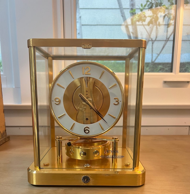 K&ouml;nnte beinhalten: Eine goldfarbene Atmos-Uhr von Jaeger-LeCoultre. Die Uhr hat ein wei&szlig;es Zifferblatt mit schwarzen r&ouml;mischen Ziffern f&uuml;r die Stunden und schwarzen Zeigern. Die Uhr ist in einer Glaskuppel eingeschlossen und steht auf einem goldfarbenen Sockel.