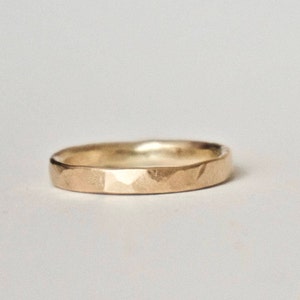 May include: A gold ring with a hammered finish. The ring features a flat band and a gently curved inner surface. The textured surface of the ring catches the light, producing highlights and shadows. A simple, elegant piece of jewellery.
